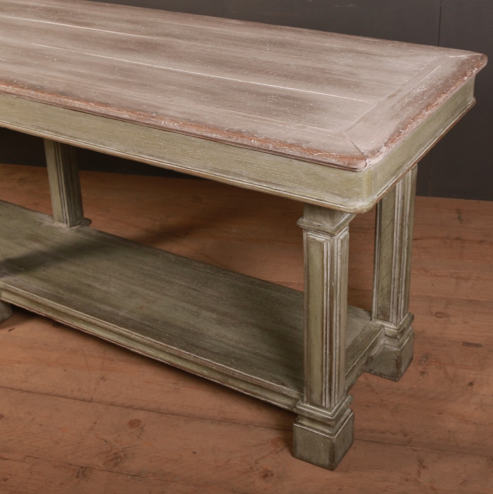 A long French drapers table featuring a muted sage green painted frame with six fluted legs, a lower full-length shelf, and a light natural timber plank top. View VII