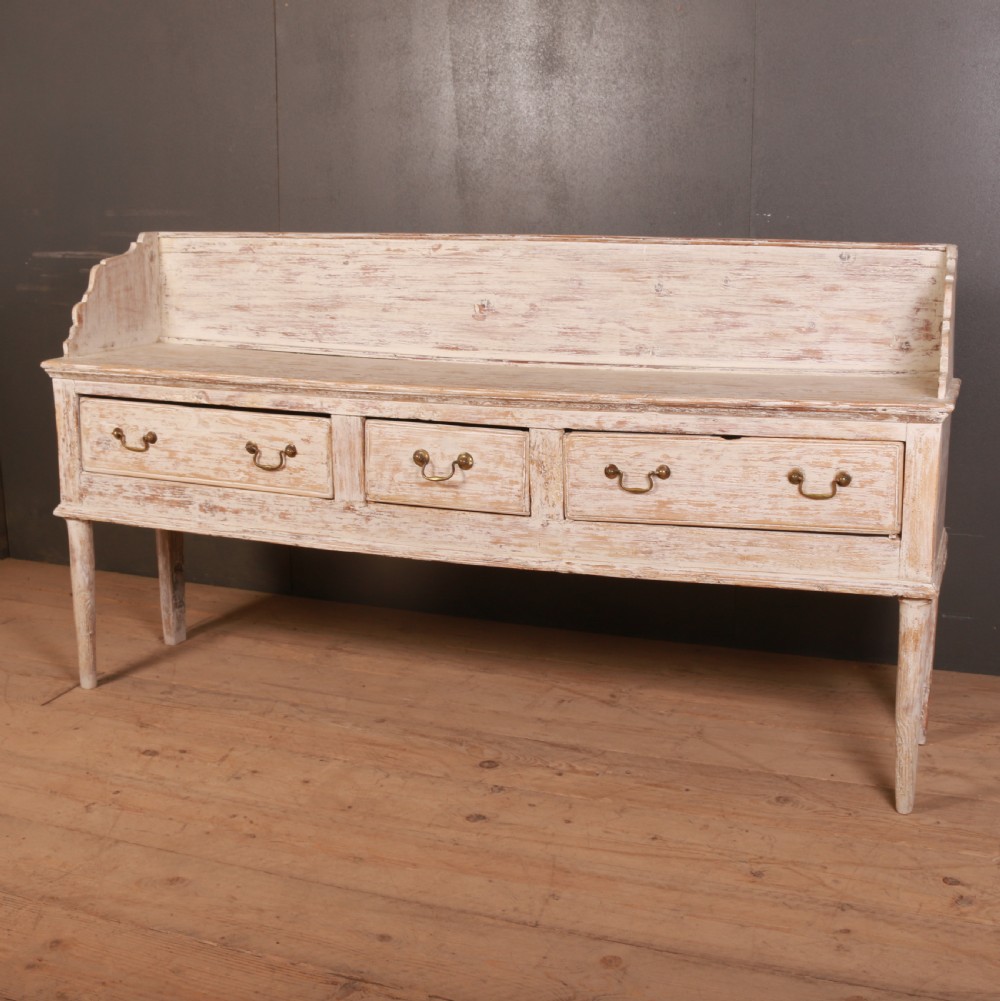 A George II painted pine dresser base with three drawers, brass drop handles, a low gallery back, and a distressed off-white finish.