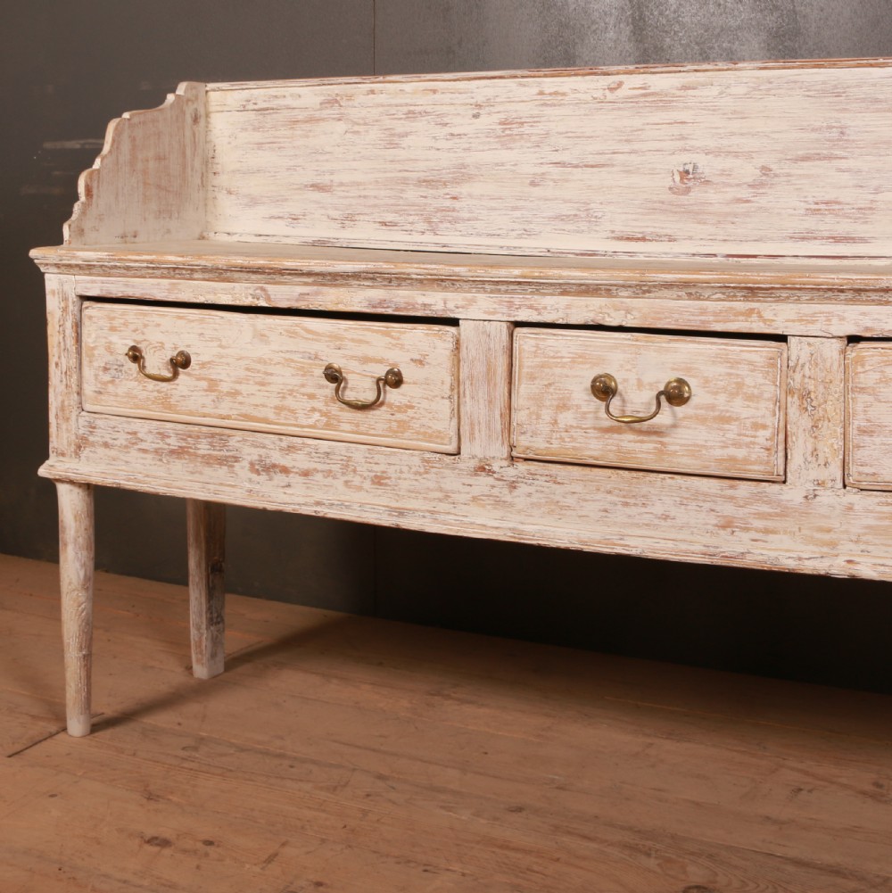 A George II painted pine dresser base with three drawers, brass drop handles, a low gallery back, and a distressed off-white finish. View II