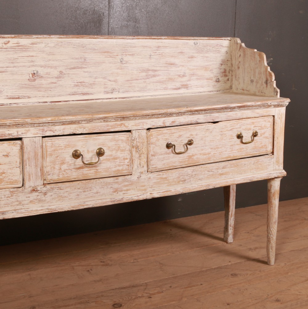 A George II painted pine dresser base with three drawers, brass drop handles, a low gallery back, and a distressed off-white finish. View IV