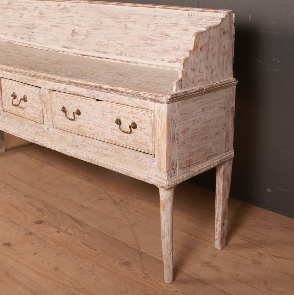 A George II painted pine dresser base with three drawers, brass drop handles, a low gallery back, and a distressed off-white finish. View V