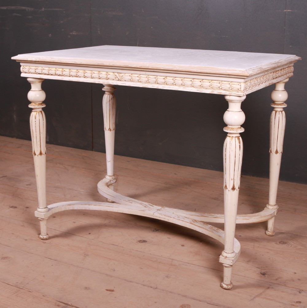 A Gustavian off-white painted lamp table featuring carved frieze detailing, gilt accents, fluted tapered legs, and a curved stretcher base.