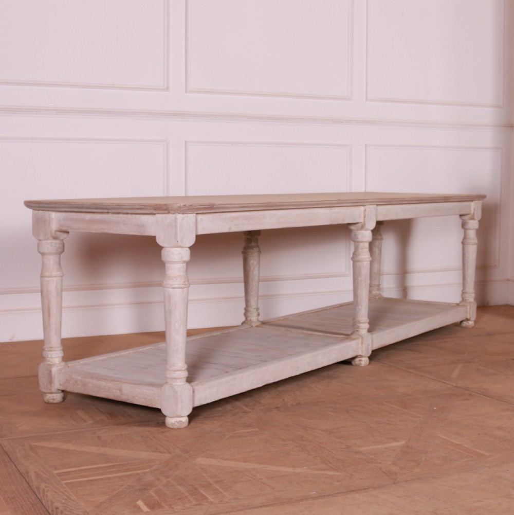 A long French painted drapers table featuring a natural wood top, six turned legs, a distressed white paint finish, and a full-length lower shelf.