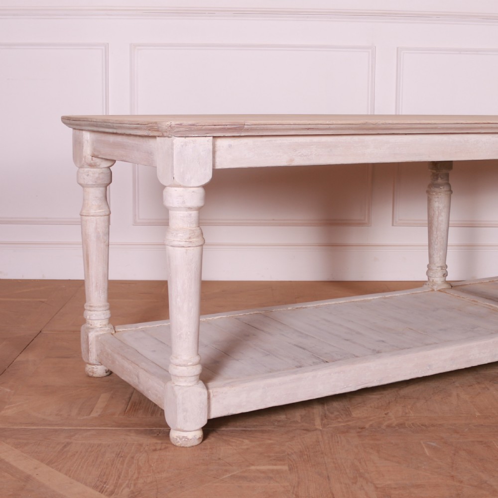 A long French painted drapers table featuring a natural wood top, six turned legs, a distressed white paint finish, and a full-length lower shelf. View II