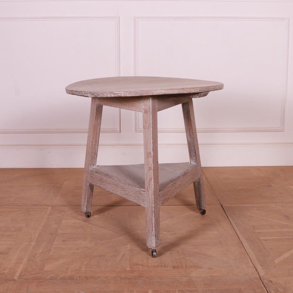 A three-legged bleached oak cricket table featuring a circular top, a triangular lower shelf, and small castors on the feet.