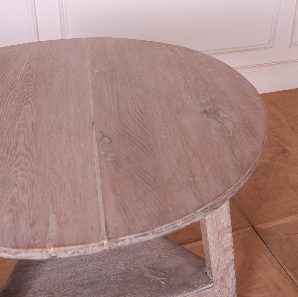A three-legged bleached oak cricket table featuring a circular top, a triangular lower shelf, and small castors on the feet. View V