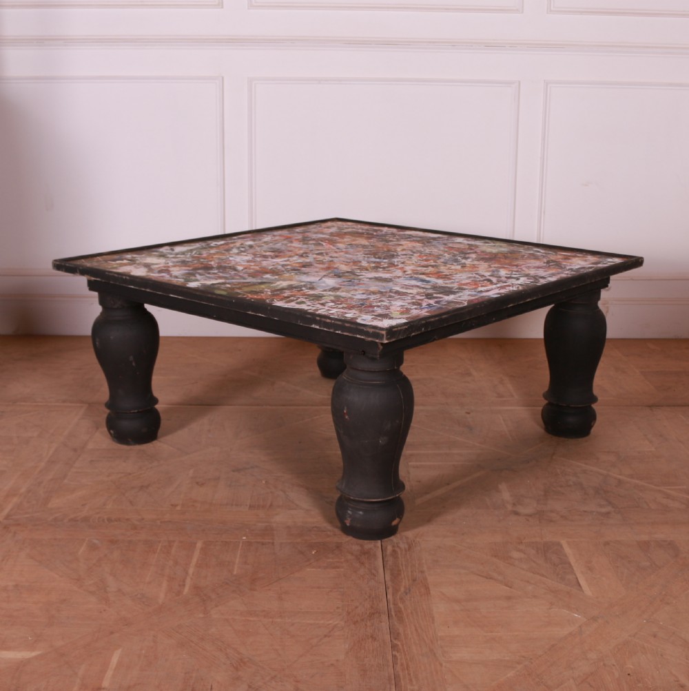 A large low square coffee table featuring heavy turned black legs and a colourful collage pattern inset into the tabletop surface.