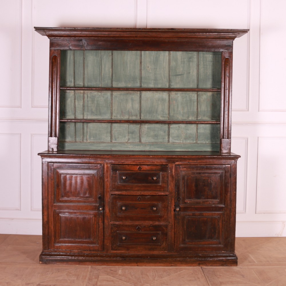 A dark pine kitchen dresser featuring an open three-tier plate rack with a sage green painted backboard above a base with three central drawers and two panelled side cupboards.
