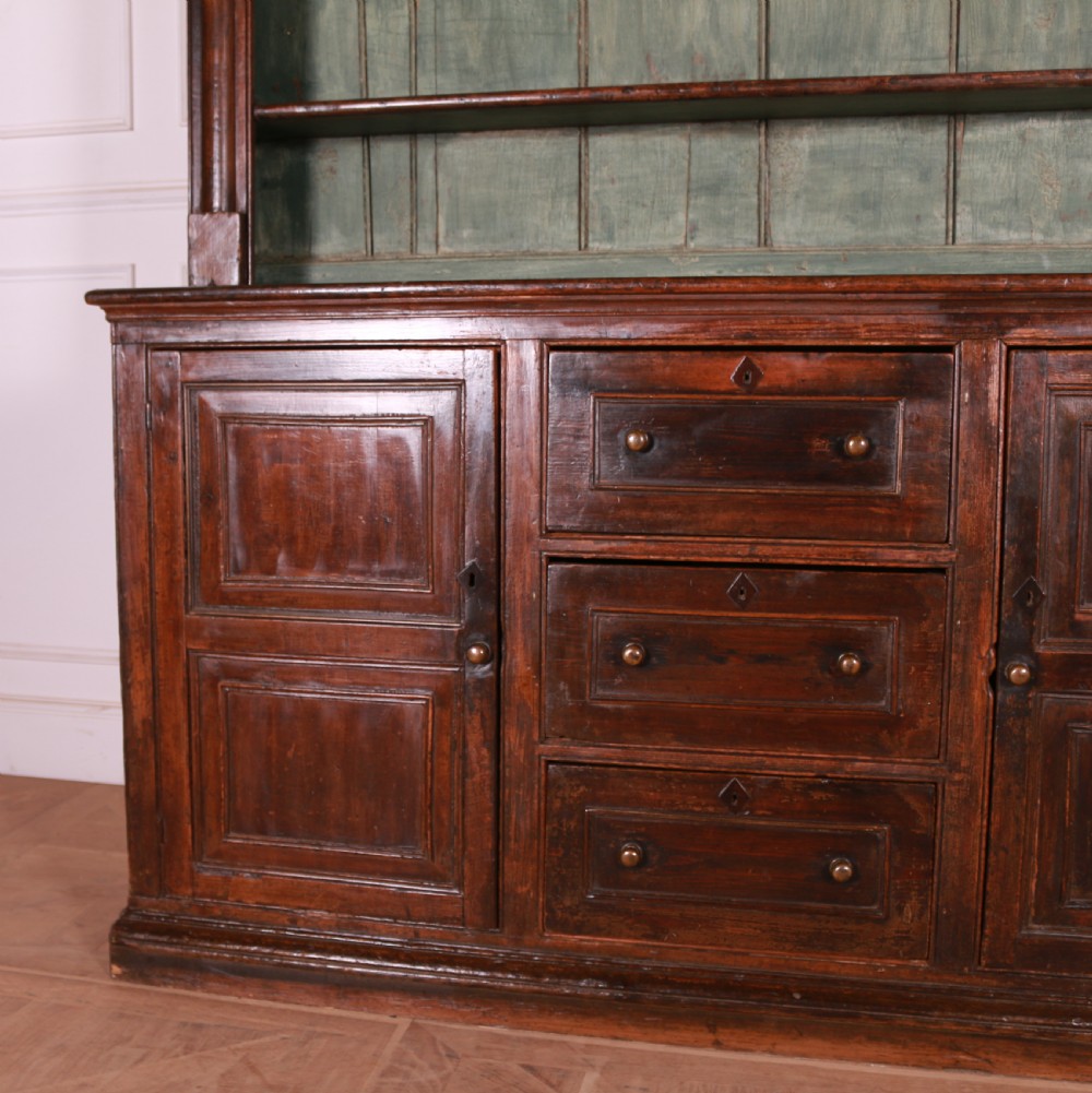 A dark pine kitchen dresser featuring an open three-tier plate rack with a sage green painted backboard above a base with three central drawers and two panelled side cupboards. View II
