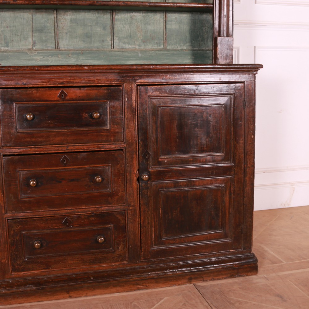 A dark pine kitchen dresser featuring an open three-tier plate rack with a sage green painted backboard above a base with three central drawers and two panelled side cupboards. View III