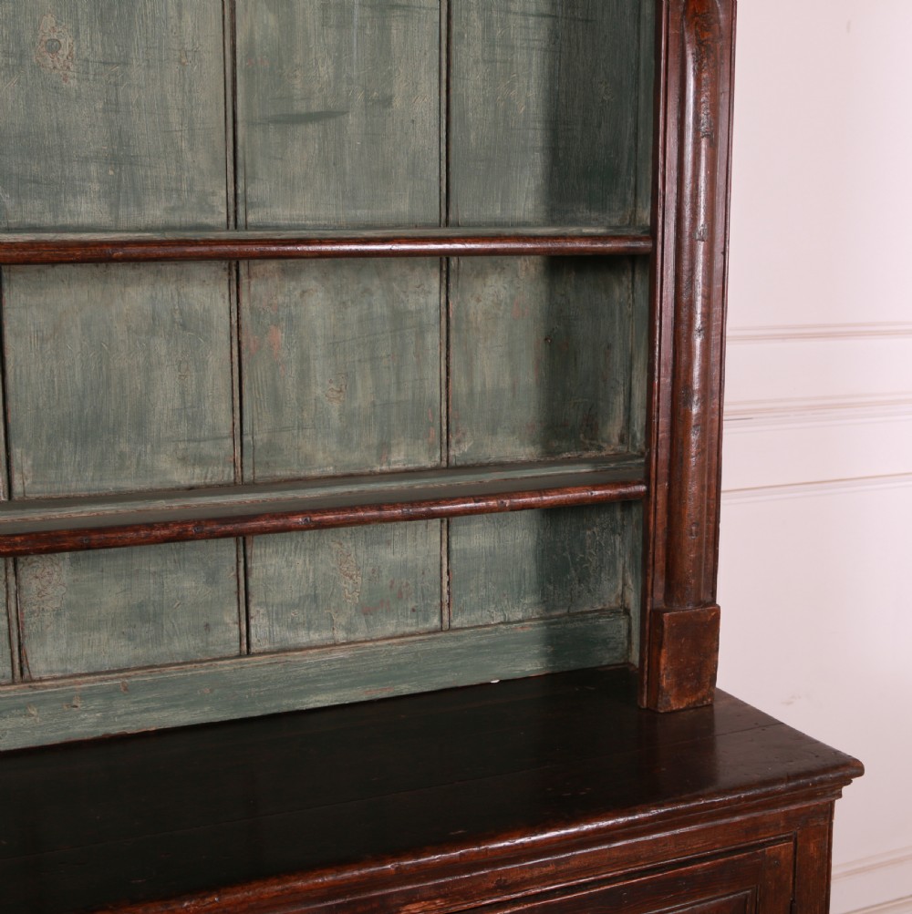 A dark pine kitchen dresser featuring an open three-tier plate rack with a sage green painted backboard above a base with three central drawers and two panelled side cupboards. View VI