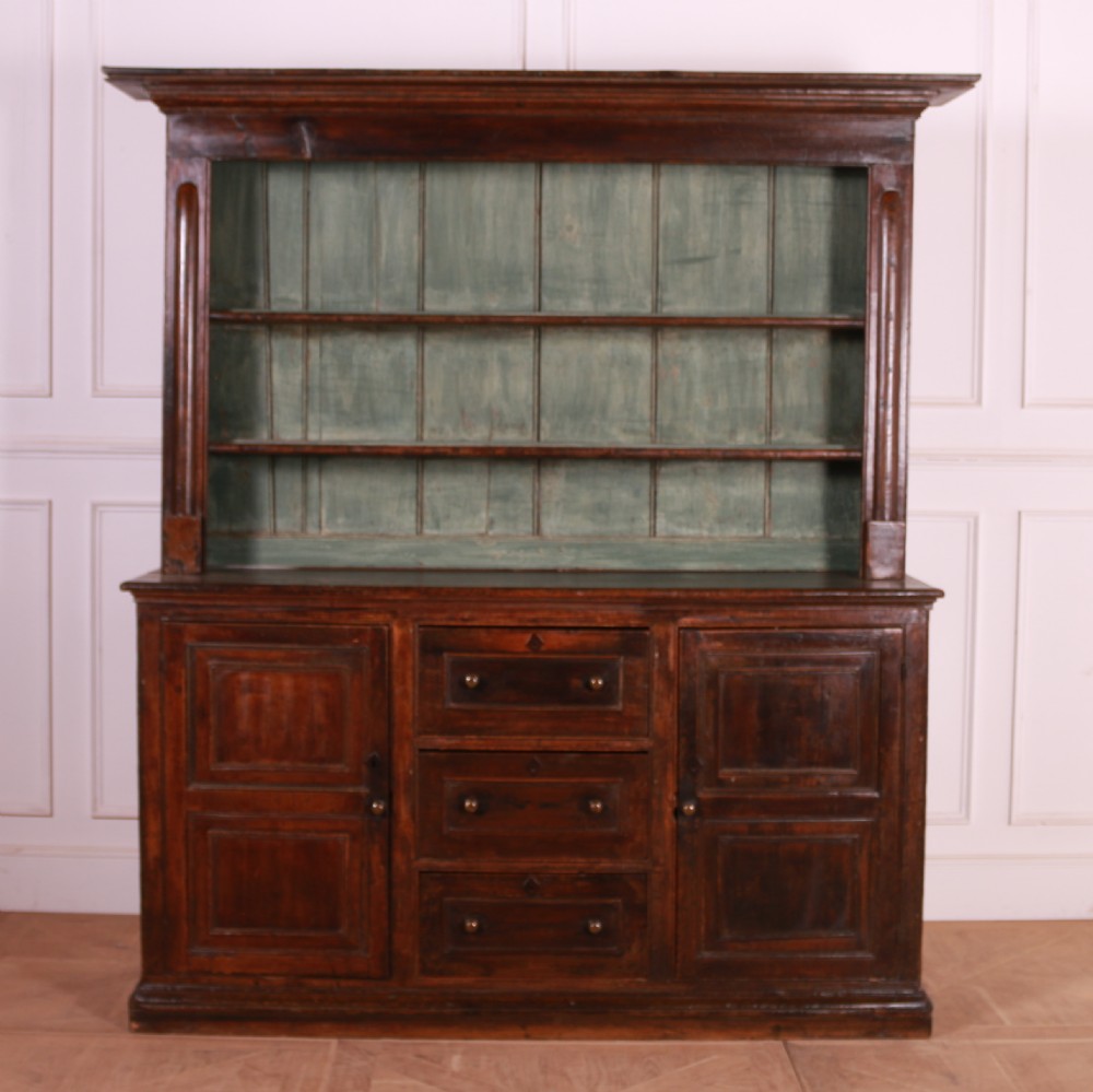 A dark pine kitchen dresser featuring an open three-tier plate rack with a sage green painted backboard above a base with three central drawers and two panelled side cupboards. View VII