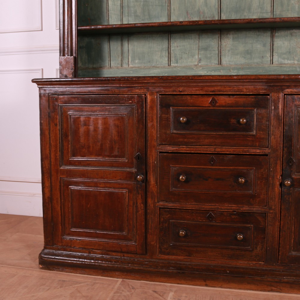 A dark pine kitchen dresser featuring an open three-tier plate rack with a sage green painted backboard above a base with three central drawers and two panelled side cupboards. View VIII