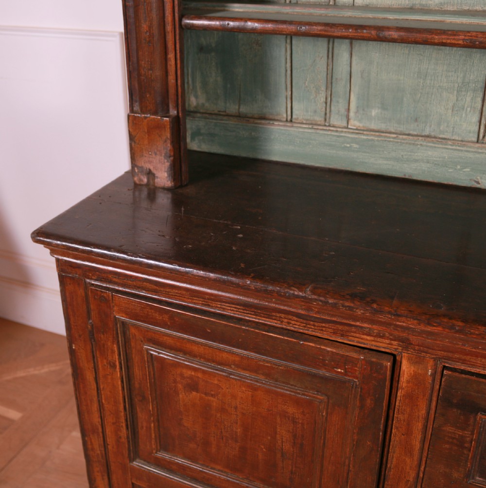 A dark pine kitchen dresser featuring an open three-tier plate rack with a sage green painted backboard above a base with three central drawers and two panelled side cupboards. View IX