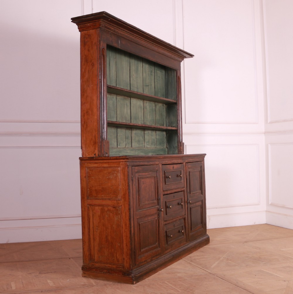 A dark pine kitchen dresser featuring an open three-tier plate rack with a sage green painted backboard above a base with three central drawers and two panelled side cupboards. View X