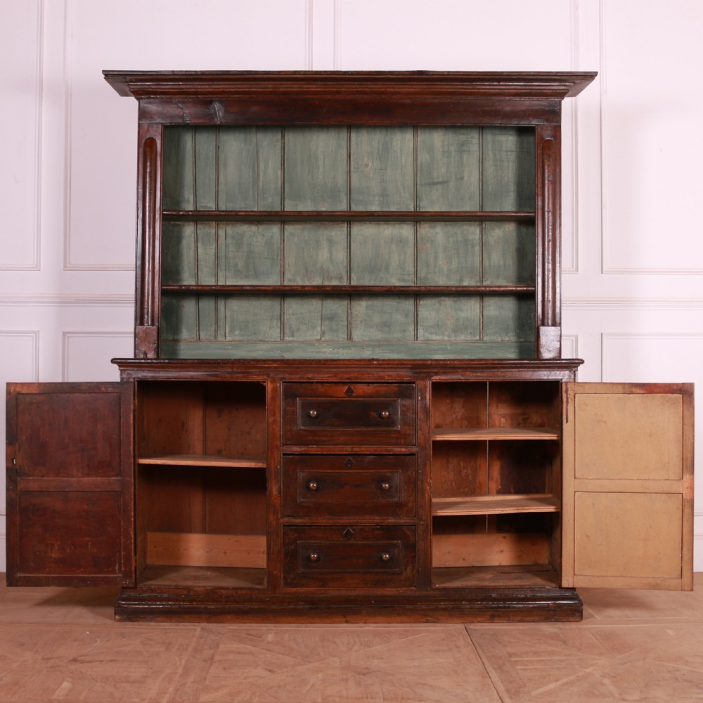 A dark pine kitchen dresser featuring an open three-tier plate rack with a sage green painted backboard above a base with three central drawers and two panelled side cupboards. View XI