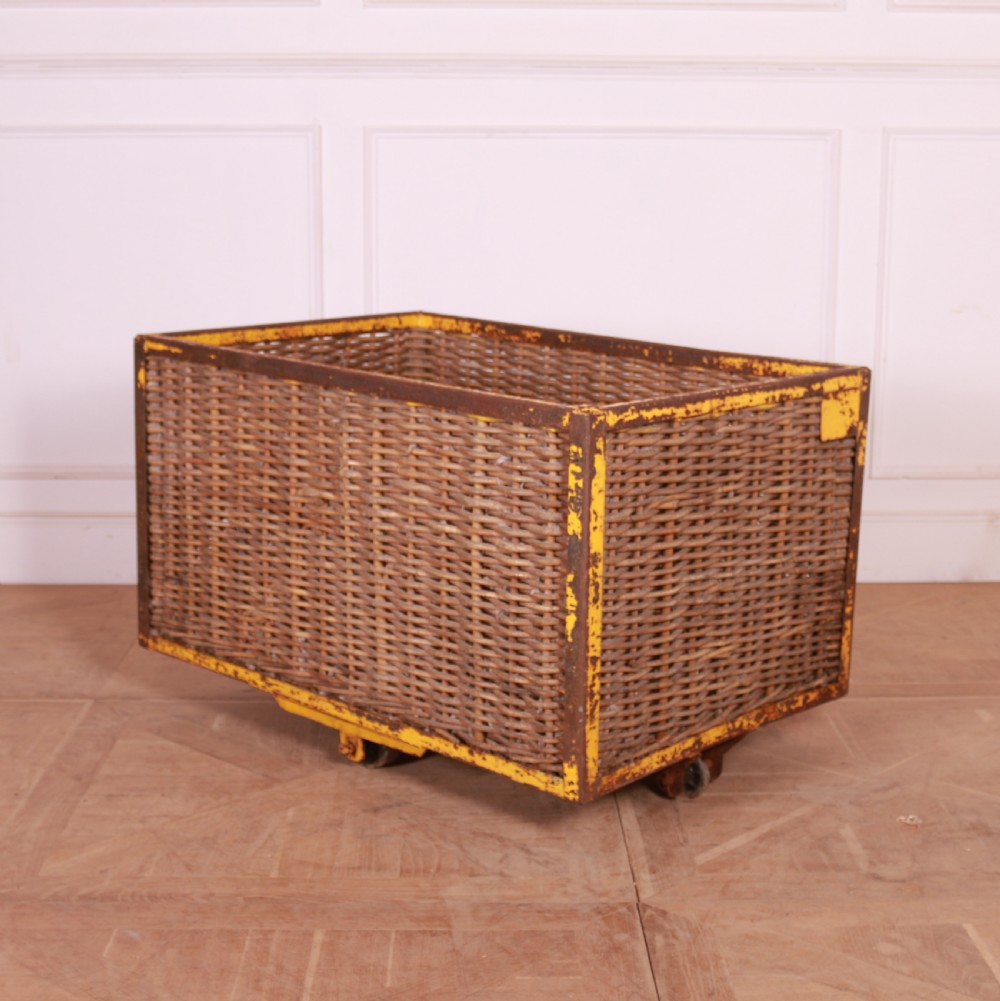 A large rectangular woven wicker basket enclosed in a distressed yellow painted steel frame with small castors on the base.