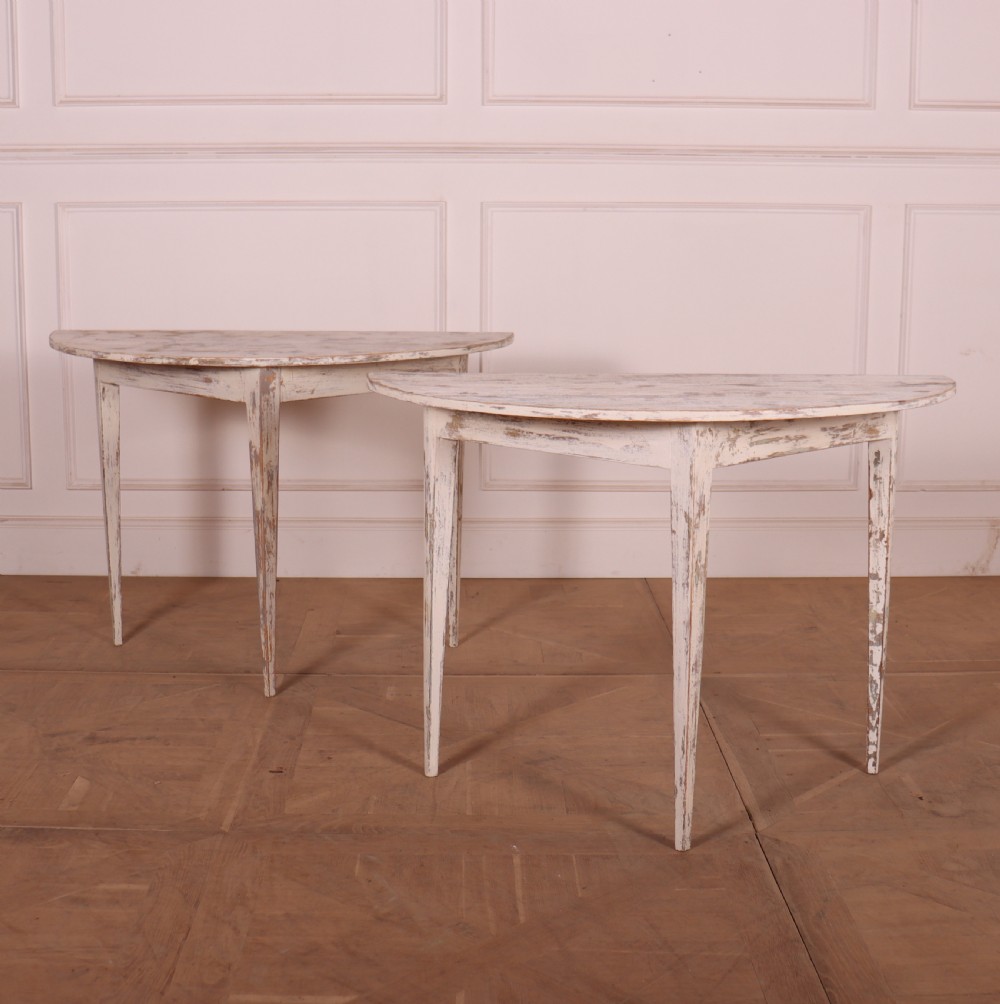 A pair of antique Swedish demi-lune console tables featuring distressed off-white painted finishes and simple tapered legs.