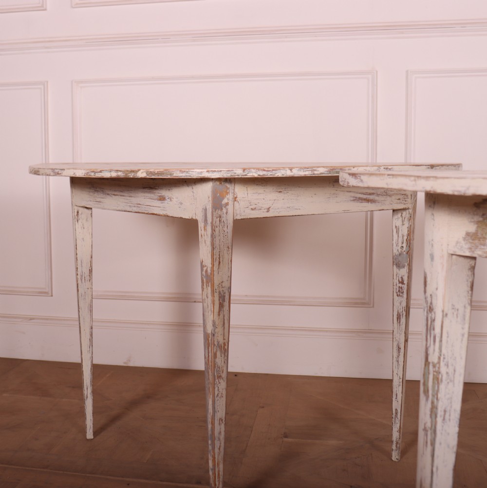 A pair of antique Swedish demi-lune console tables featuring distressed off-white painted finishes and simple tapered legs. View III