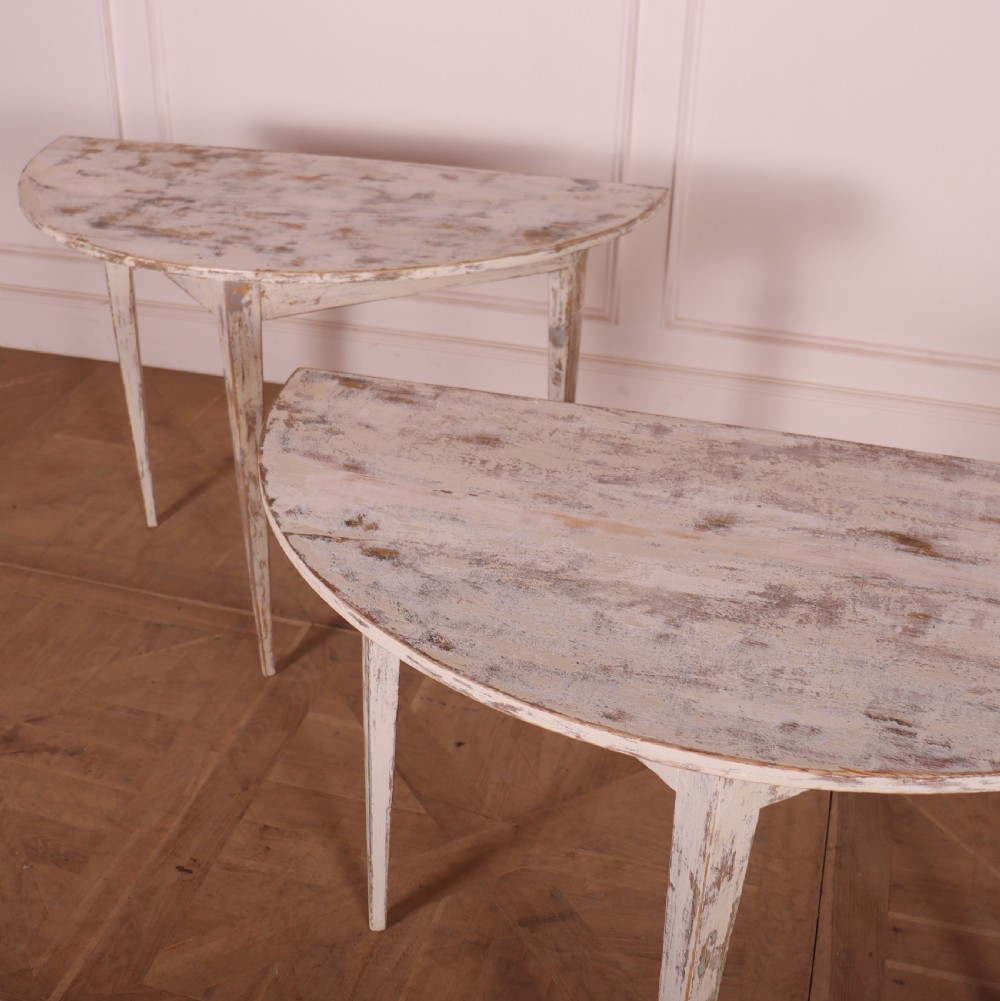 A pair of antique Swedish demi-lune console tables featuring distressed off-white painted finishes and simple tapered legs. View IV