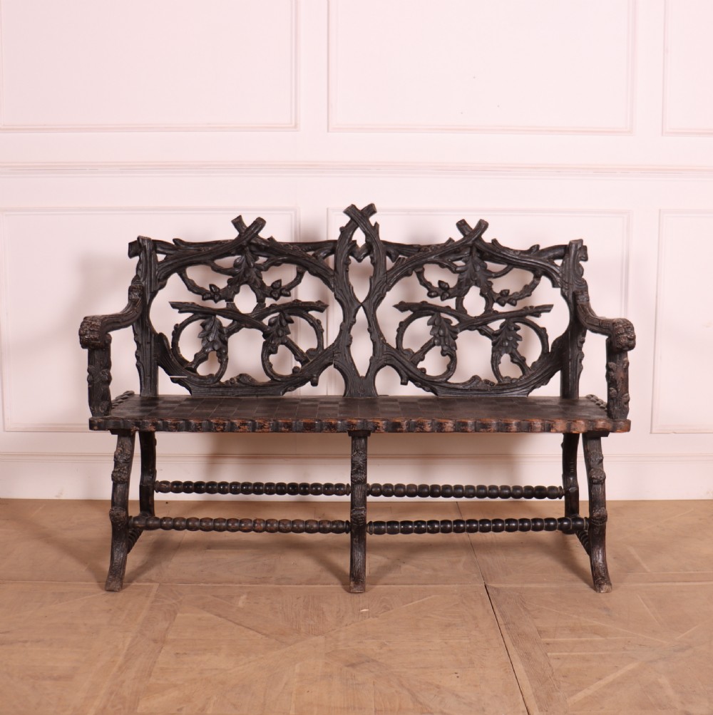 A dark wood Victorian bench with intricate branch and leaf carvings on the backrest, carved armrests, a textured seat, and bobbin stretchers.