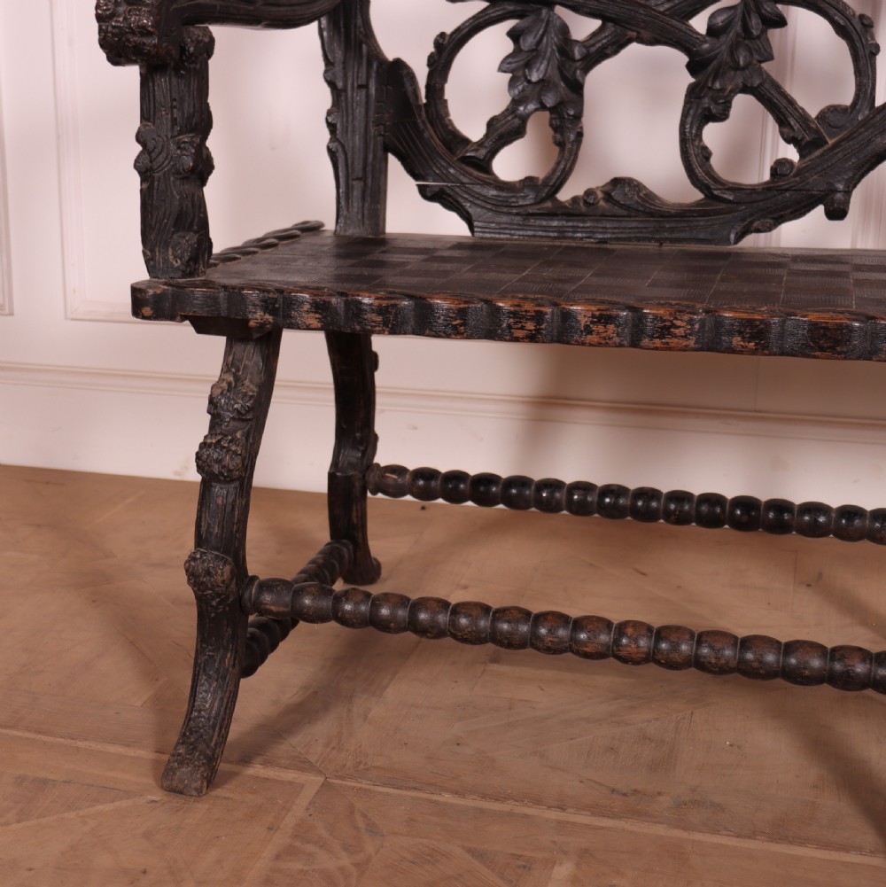 A dark wood Victorian bench with intricate branch and leaf carvings on the backrest, carved armrests, a textured seat, and bobbin stretchers. View IV
