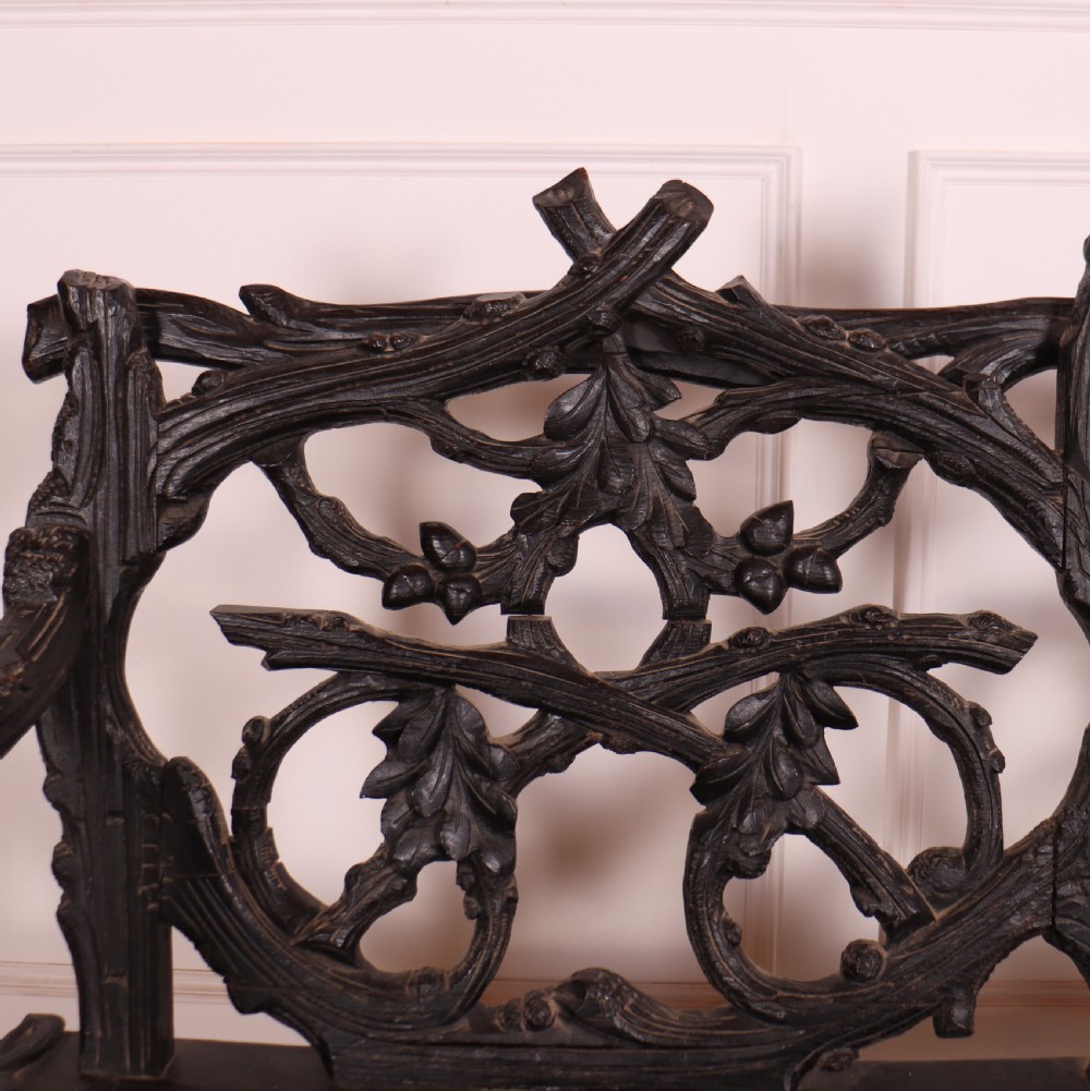 A dark wood Victorian bench with intricate branch and leaf carvings on the backrest, carved armrests, a textured seat, and bobbin stretchers. View V