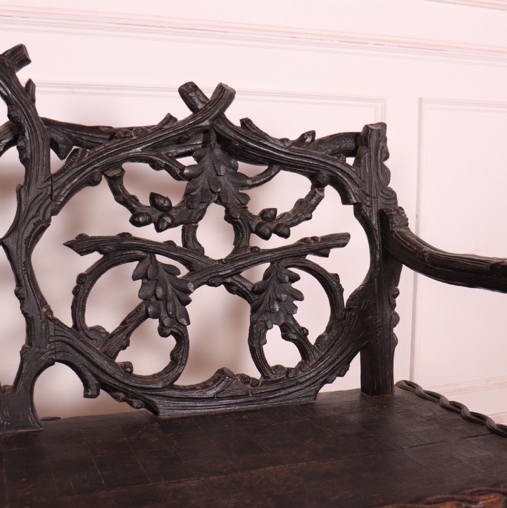 A dark wood Victorian bench with intricate branch and leaf carvings on the backrest, carved armrests, a textured seat, and bobbin stretchers. View VI