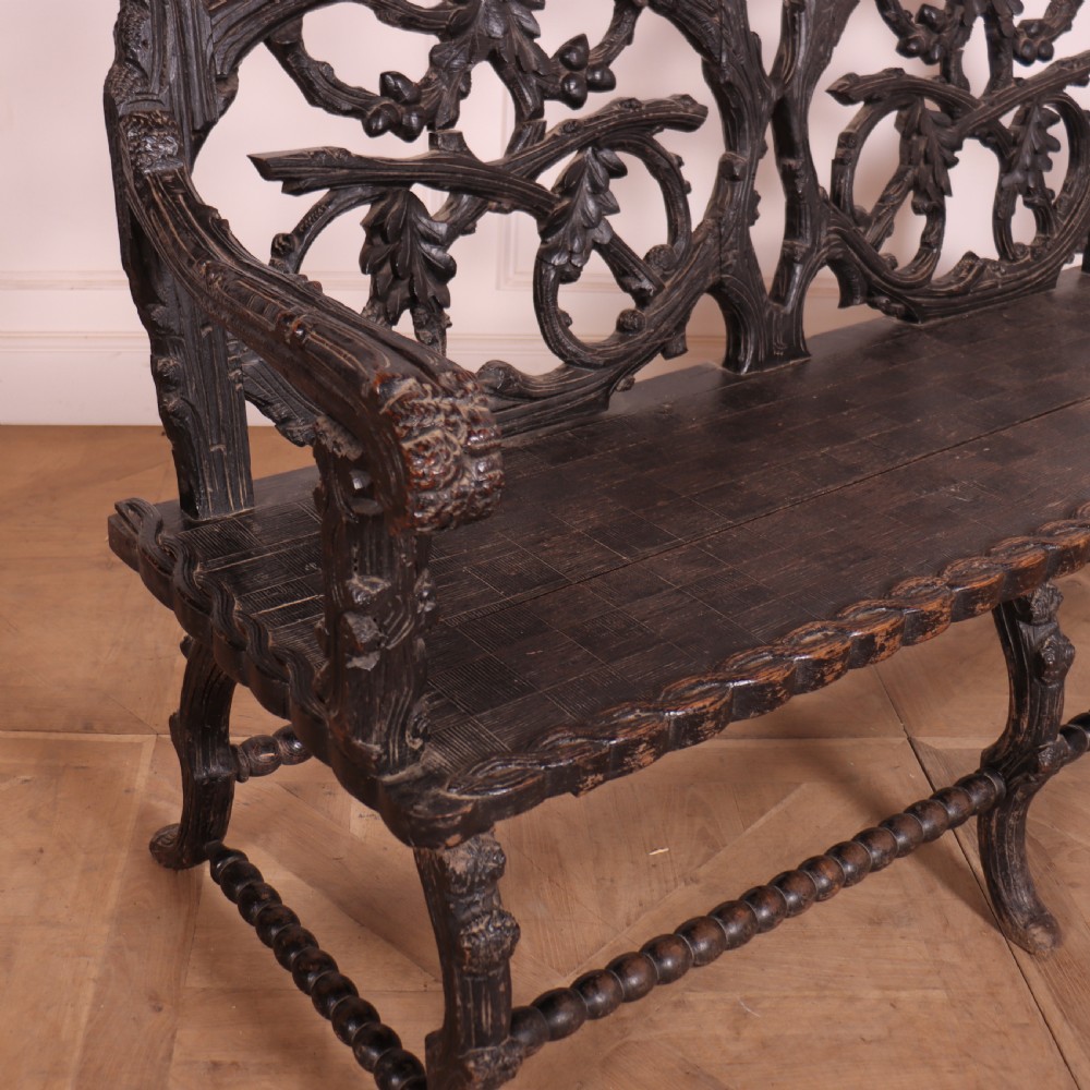 A dark wood Victorian bench with intricate branch and leaf carvings on the backrest, carved armrests, a textured seat, and bobbin stretchers. View VII
