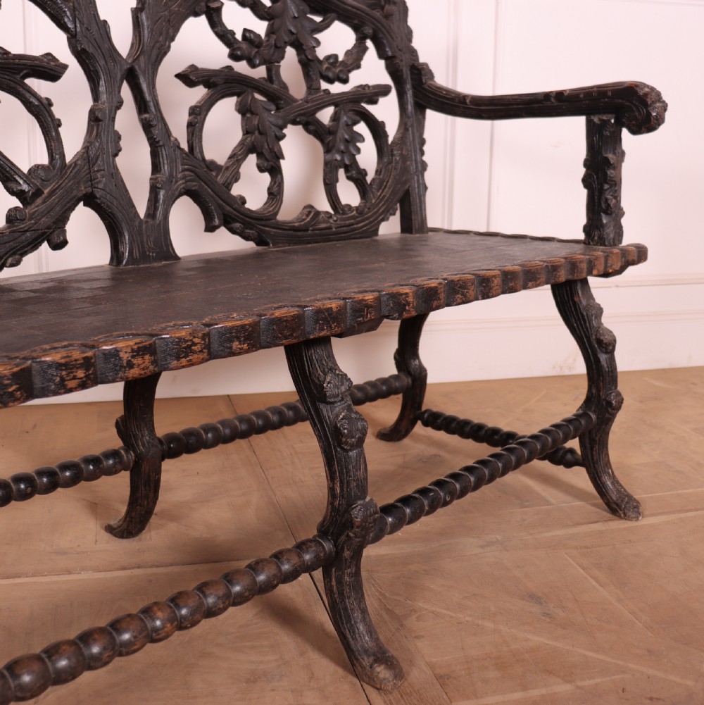A dark wood Victorian bench with intricate branch and leaf carvings on the backrest, carved armrests, a textured seat, and bobbin stretchers. View IX