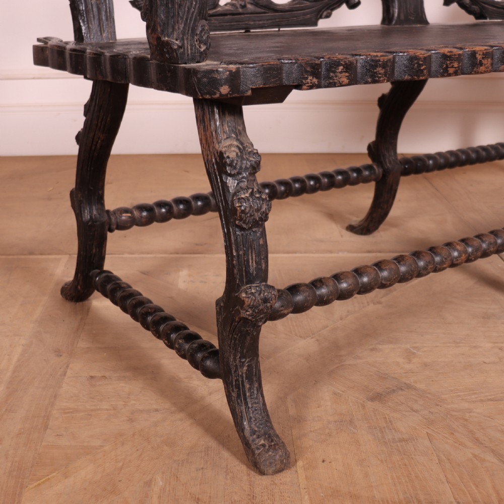 A dark wood Victorian bench with intricate branch and leaf carvings on the backrest, carved armrests, a textured seat, and bobbin stretchers. View X