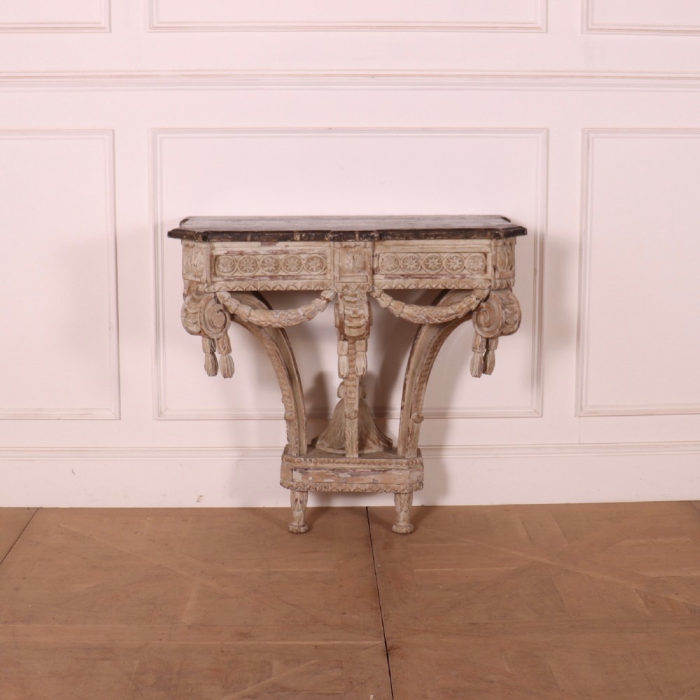 An 18th-century French carved wood console table featuring a dark top, rosette frieze, carved drapery swags, and a distressed off-white finish.