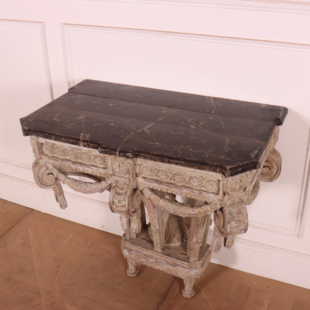 An 18th-century French carved wood console table featuring a dark top, rosette frieze, carved drapery swags, and a distressed off-white finish. View V