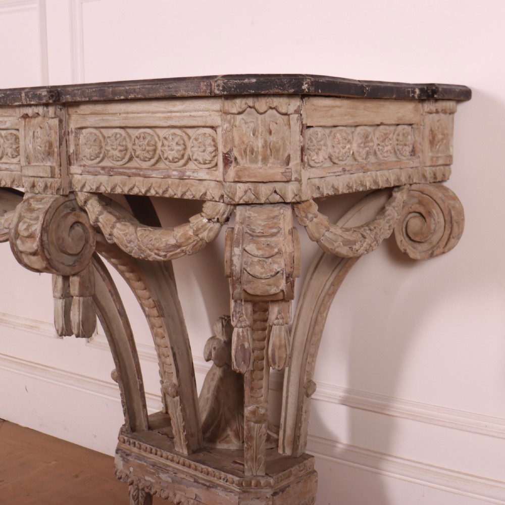 An 18th-century French carved wood console table featuring a dark top, rosette frieze, carved drapery swags, and a distressed off-white finish. View VI