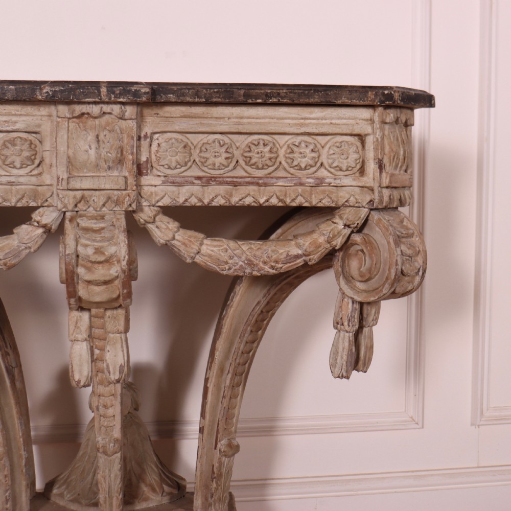 An 18th-century French carved wood console table featuring a dark top, rosette frieze, carved drapery swags, and a distressed off-white finish. View VII