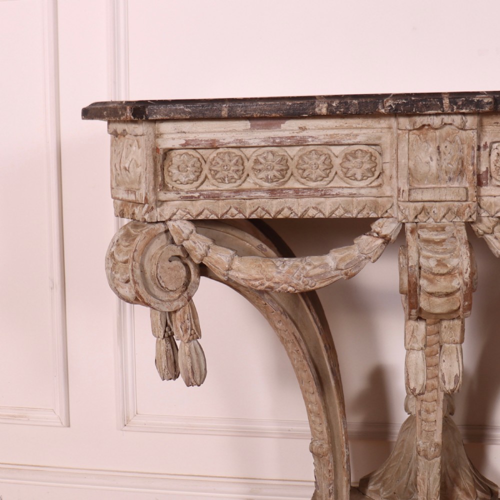 An 18th-century French carved wood console table featuring a dark top, rosette frieze, carved drapery swags, and a distressed off-white finish. View IX