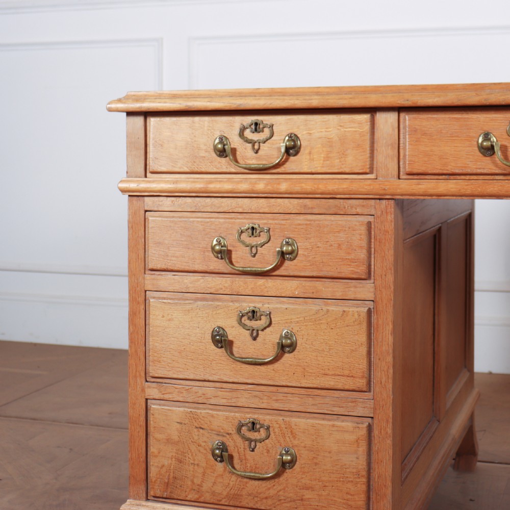 An antique pale oak pedestal desk featuring ten drawers with brass drop handles and a dark leather inset writing surface. View V