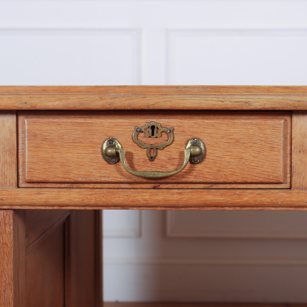 An antique pale oak pedestal desk featuring ten drawers with brass drop handles and a dark leather inset writing surface. View VI