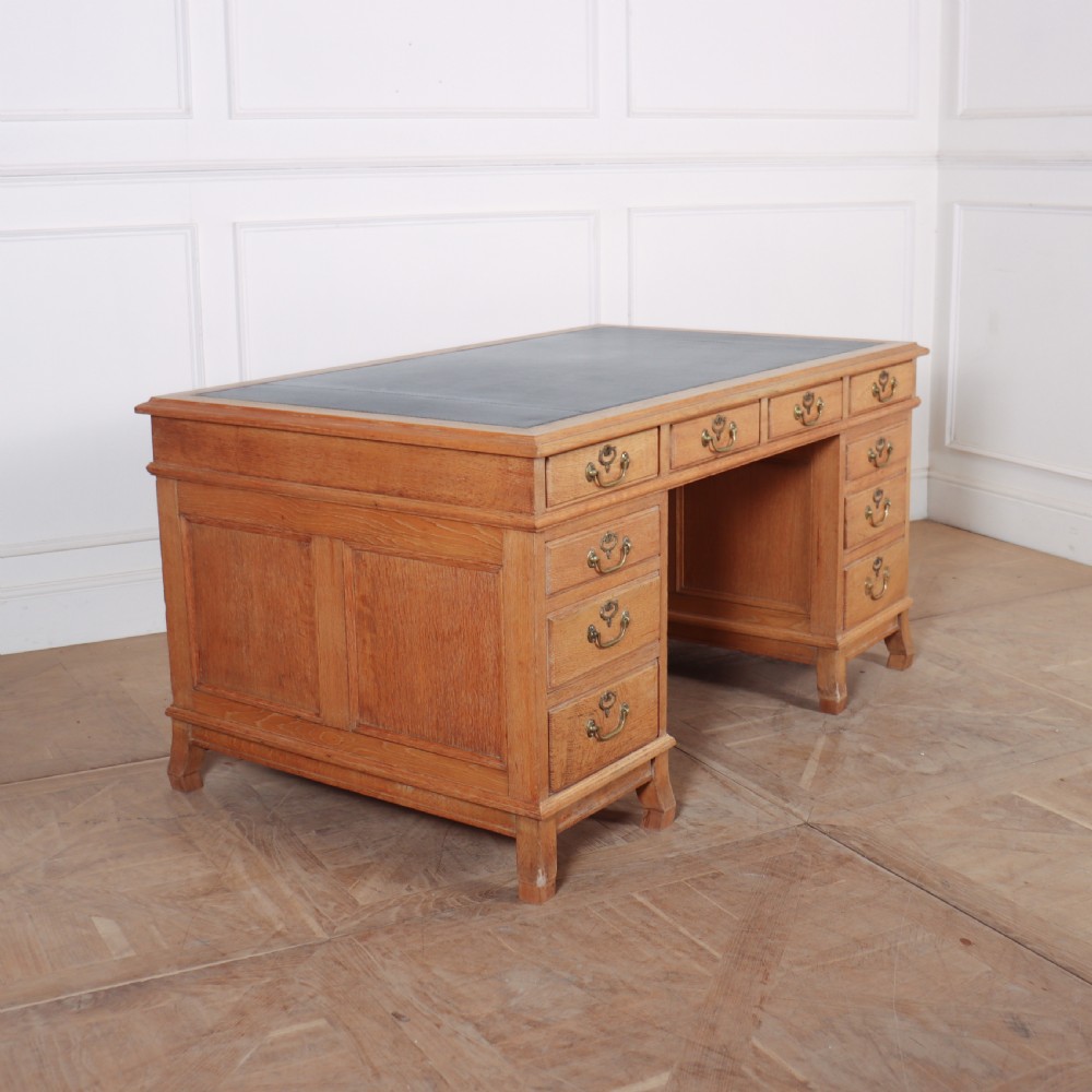 An antique pale oak pedestal desk featuring ten drawers with brass drop handles and a dark leather inset writing surface. View VII