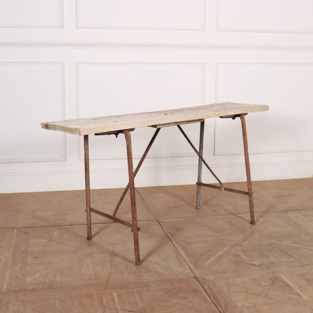 A long, narrow bleached timber top resting on a slender, oxidised metal trestle frame.