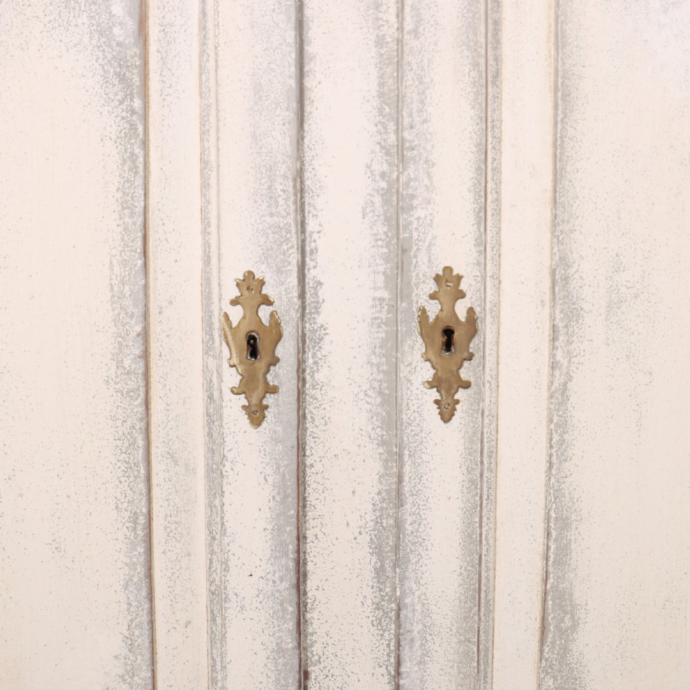 An 18th-century Swedish painted wooden linen cupboard featuring panelled double doors, brass keyhole escutcheons, and a distressed off-white painted finish. View VI