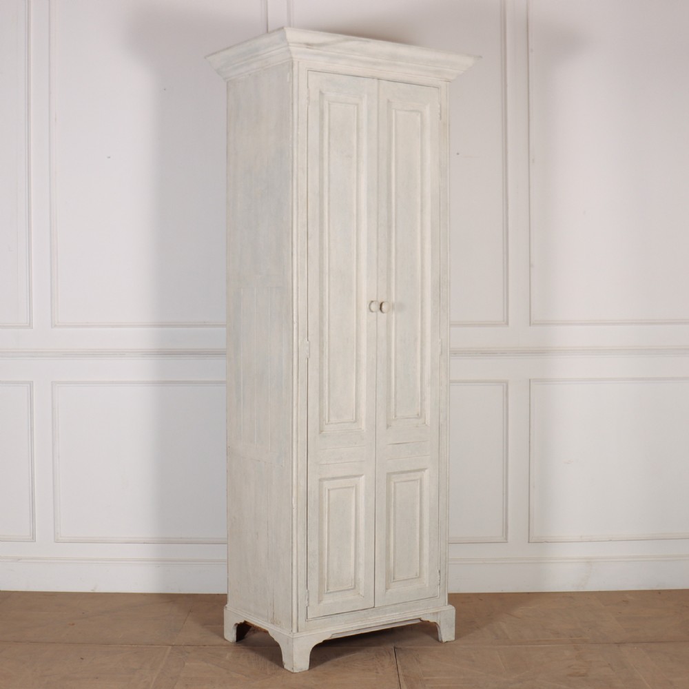 A tall, narrow antique off-white painted wooden cupboard featuring double panelled doors, small central wooden knobs, a simple moulded top cornice, and bracket feet.