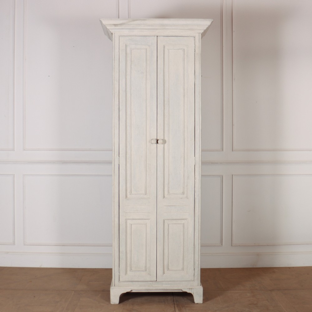 A tall, narrow antique off-white painted wooden cupboard featuring double panelled doors, small central wooden knobs, a simple moulded top cornice, and bracket feet. View II
