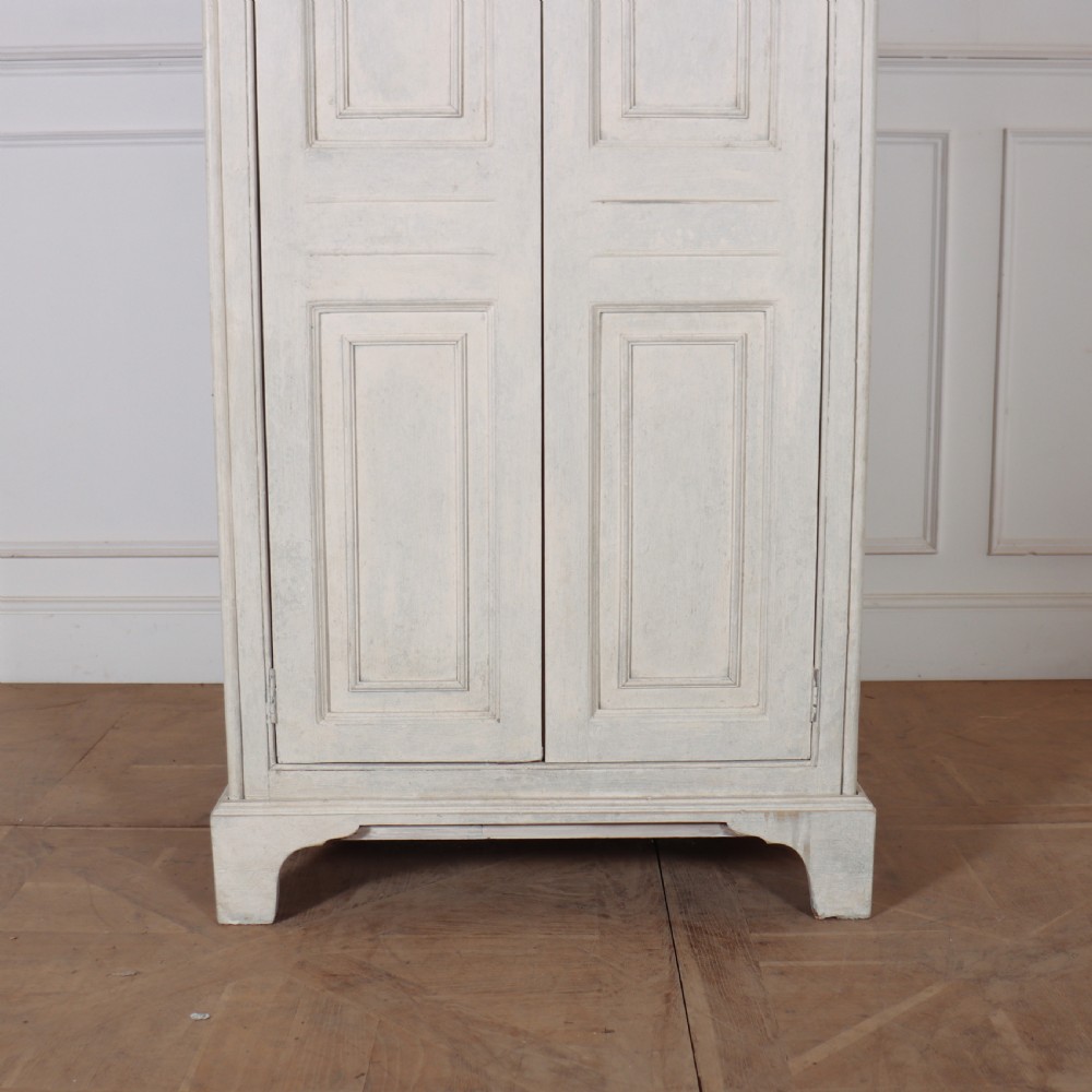 A tall, narrow antique off-white painted wooden cupboard featuring double panelled doors, small central wooden knobs, a simple moulded top cornice, and bracket feet. View III