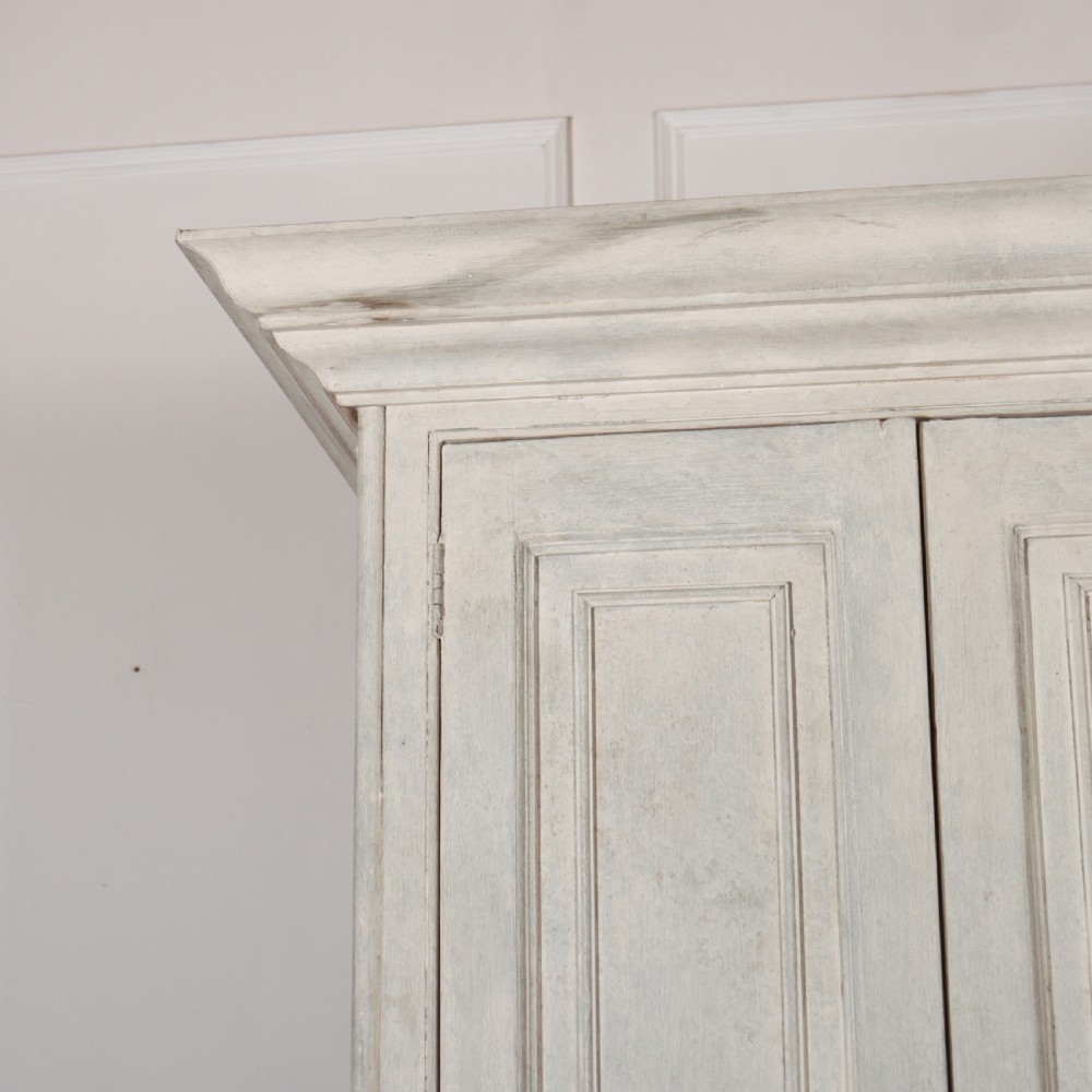 A tall, narrow antique off-white painted wooden cupboard featuring double panelled doors, small central wooden knobs, a simple moulded top cornice, and bracket feet. View IV