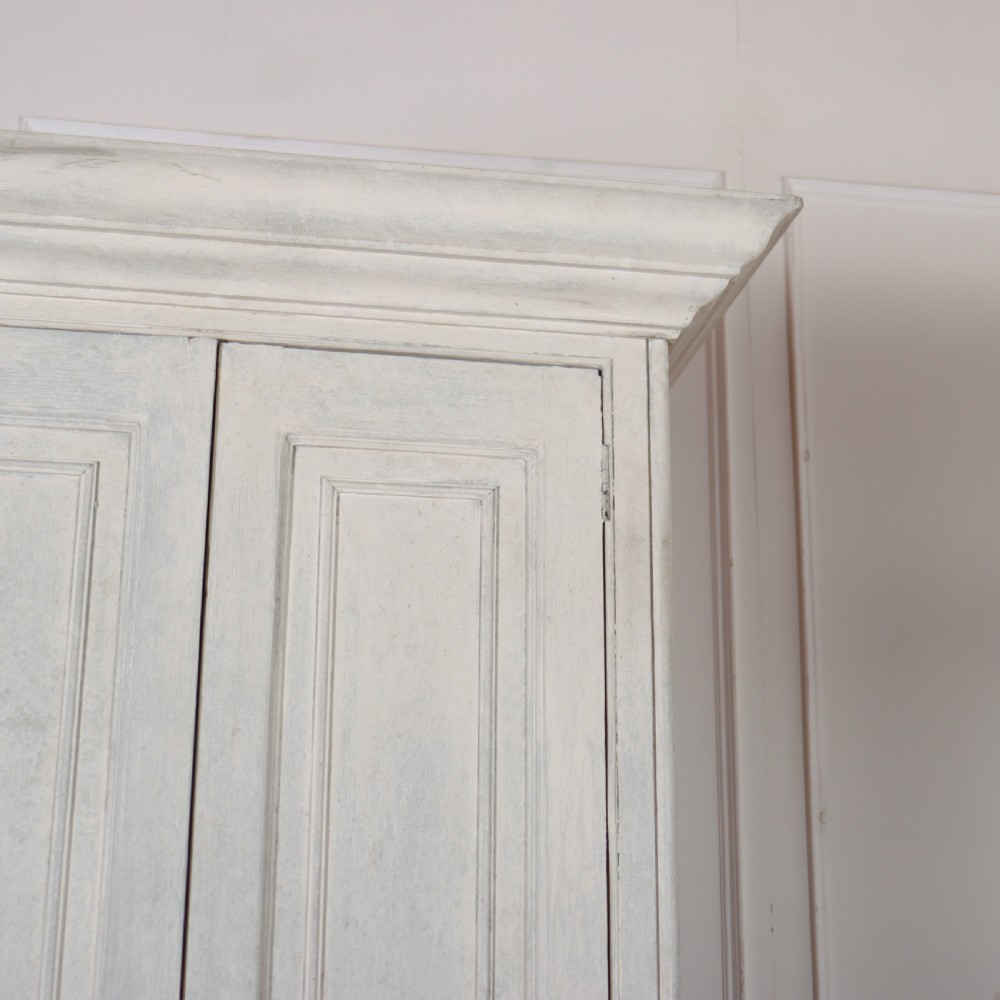 A tall, narrow antique off-white painted wooden cupboard featuring double panelled doors, small central wooden knobs, a simple moulded top cornice, and bracket feet. View V