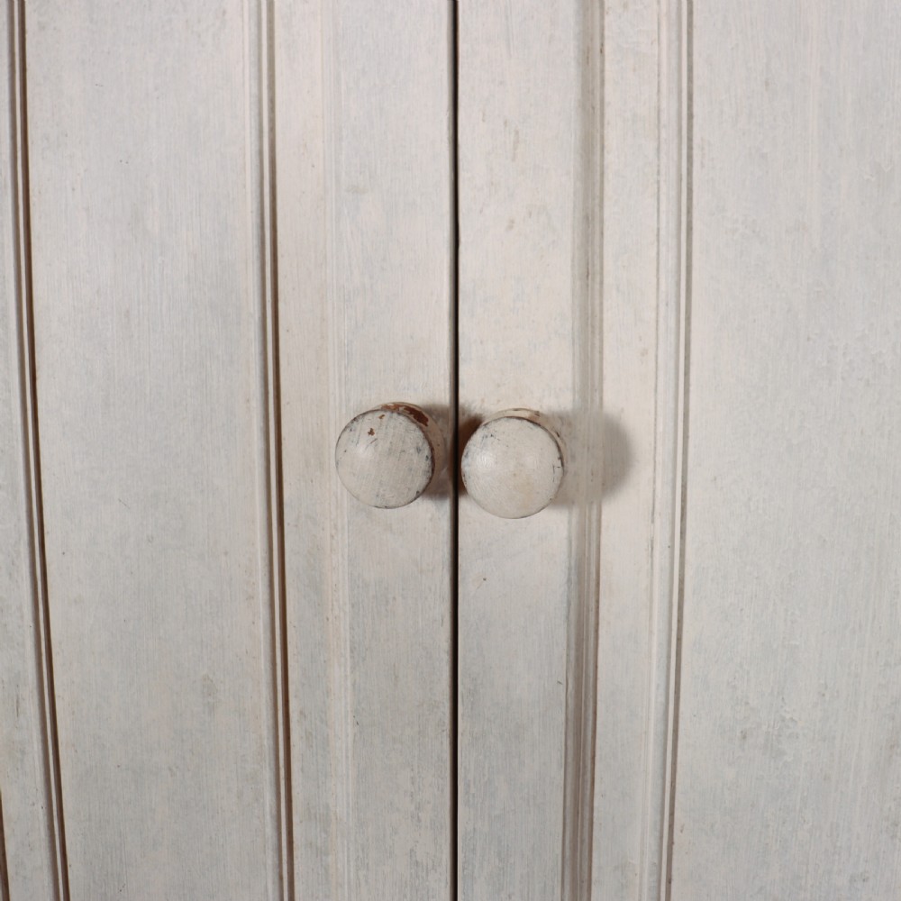A tall, narrow antique off-white painted wooden cupboard featuring double panelled doors, small central wooden knobs, a simple moulded top cornice, and bracket feet. View VI