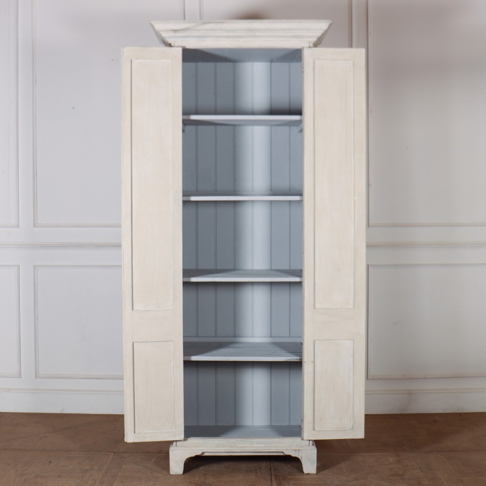 A tall, narrow antique off-white painted wooden cupboard featuring double panelled doors, small central wooden knobs, a simple moulded top cornice, and bracket feet. View VII