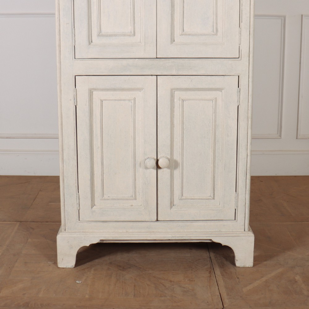 A tall, narrow off-white painted pine linen cupboard featuring two upper panelled doors and two lower panelled doors with turned wooden knobs. View V