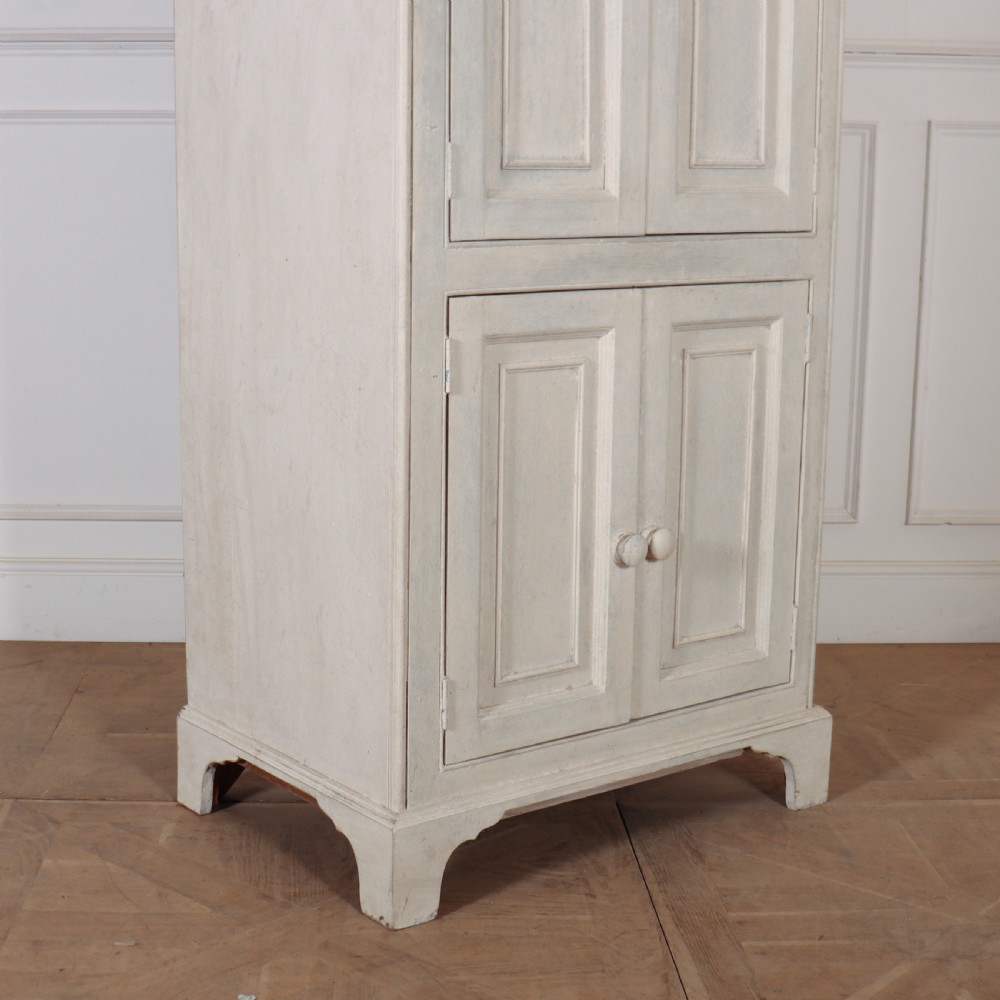 A tall, narrow off-white painted pine linen cupboard featuring two upper panelled doors and two lower panelled doors with turned wooden knobs. View VI